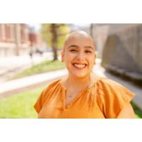 Natalie's headshot, standing outside on a sunny day in an orange top and smiling.